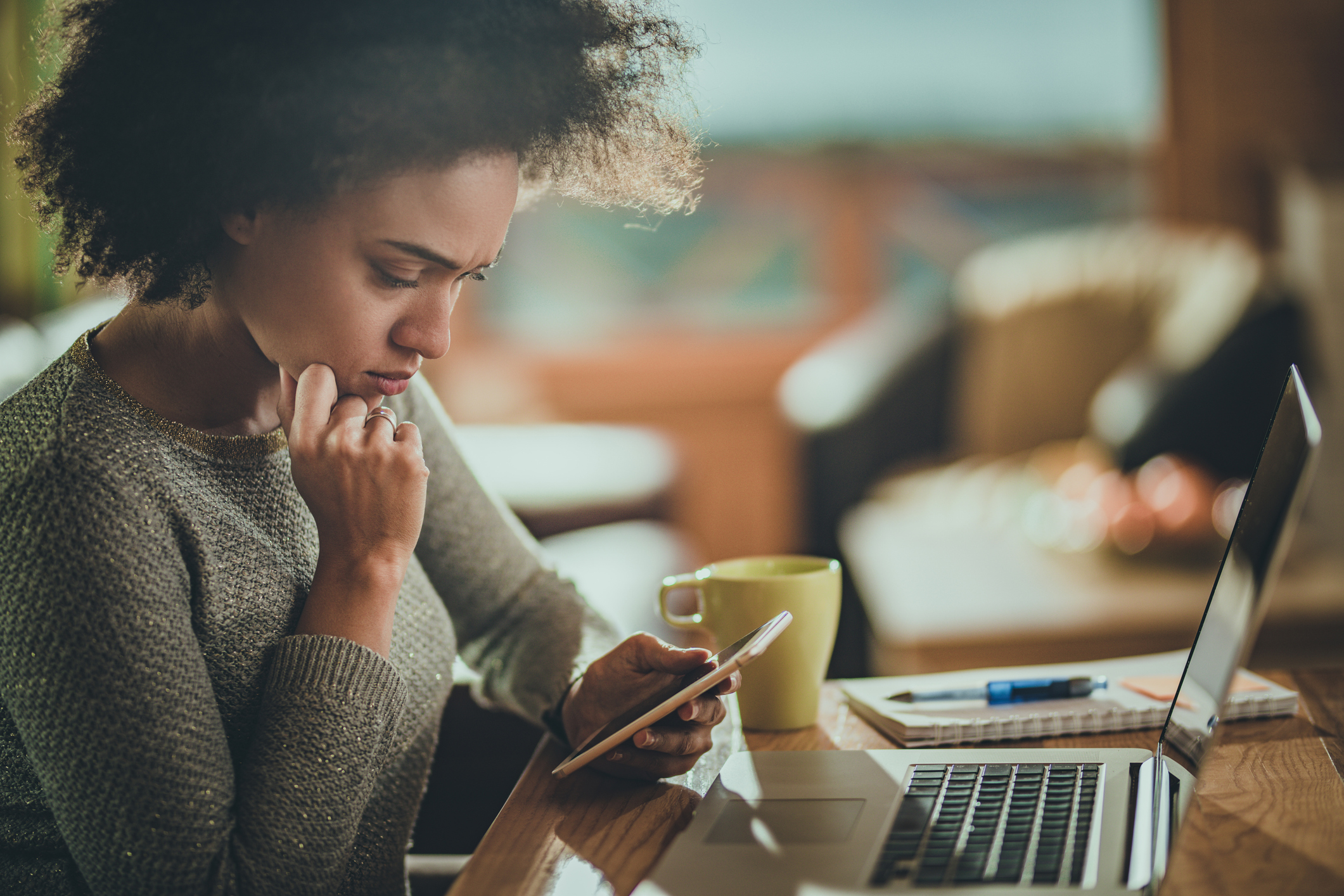 Woman sitting at an open laptop looking at her phone with concern